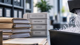 Empilements de documents de bureau à côté de boîtes d’archives étiquetées ressources humaines, comptabilité, contrats et archives, avec une déchiqueteuse au premier plan